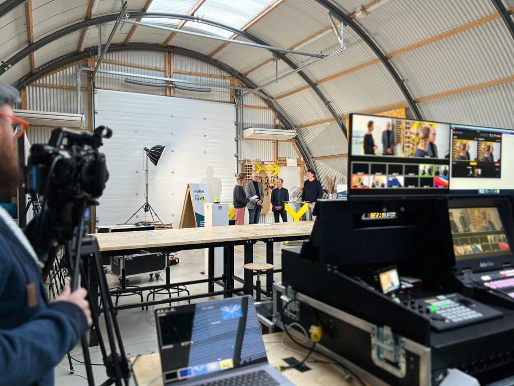 Opname van een certificeringsmoment bij Cibap Next. In een werkplaatsomgeving worden sprekers gefilmd terwijl ze een presentatie geven, met een cameraman en een videoregie op de voorgrond. Op de schermen is de live-registratie te zien.