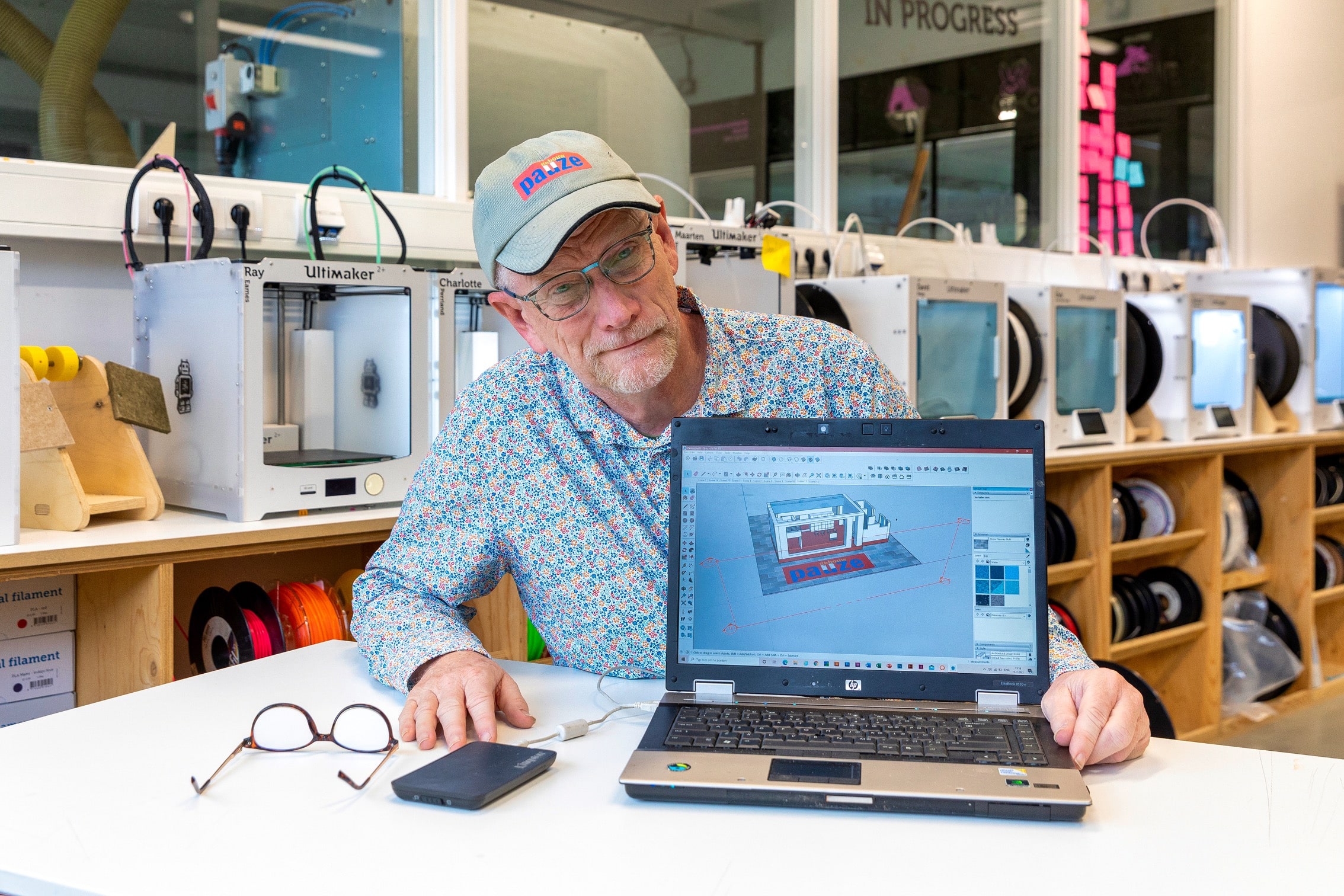 Een man zit achter een computer waarop hij een 3D-ontwerp van een woning toont in SketchUp Pro. Op de achtergrond staan 3D-printers in Cibap vakschool