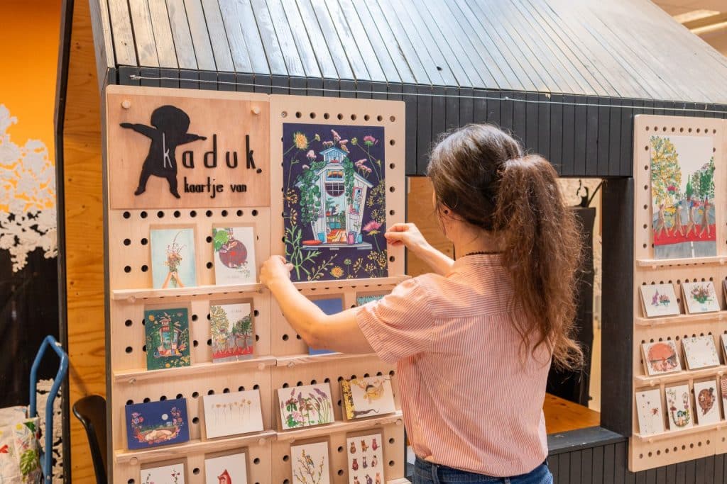 Katja Kaduk, deelnemer van de training meubelmaken basis van Cibap Next, naast haar zelf ontworpen meubelstuk van hout: een modulair kaartenrek voor haar illustraties. Ze houdt een illustratie vast die ze in het rek zet.