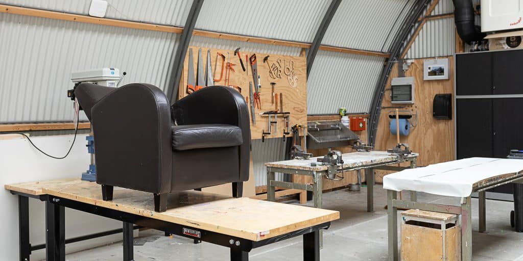 Werkplaats voor de opleiding tot meubelreparateur bij Cibap Next. Een leren fauteuil staat op een werkbank, omringd door gereedschappen en werktafels, klaar voor restauratie. De ruimte is ingericht voor vakmanschap en ambachtelijke meubelreparatie.